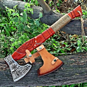 May include: A handcrafted axe with a wooden handle wrapped in twine and a leather sheath. The axe head is silver with etched designs. The handle is a reddish-brown colour. The axe is resting on a tree branch.
