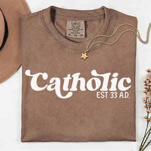 May include: A brown t-shirt with the word "Catholic" in white script and "EST 33 A.D." printed below. A gold star pendant necklace rests on the shirt. A brown hat and flowers are in the background.