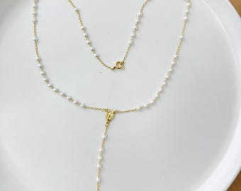 Puede incluir: Un collar de rosario dorado con pequeñas cuentas blancas y un colgante de cruz. El collar se exhibe sobre un plato blanco, mostrando su delicado diseño y significado religioso. La cadena y la cruz del rosario son doradas.
