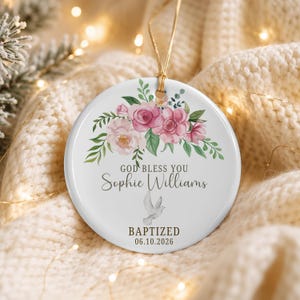 Pode incluir: Um ornamento redondo de cerâmica branca com uma ilustração floral de rosas cor-de-rosa e folhas verdes no topo. O texto diz GOD BLESS YOU Sophie Williams BAPTIZED 06.10.2026 com uma pequena pomba cinzenta e um cordão dourado.