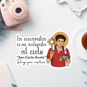 Puede incluir: Una pegatina con una ilustración de un joven sosteniendo una custodia y un mando de videojuegos. El texto dice "La eucaristia es mi autopista al cielo" y "San Carlo Acutis". El hombre lleva una camisa polo roja.