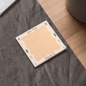 May include: A square notepad with a light peach center and a border featuring religious icons. The text "Virgin Mary" and "Pray for Us" are written in a script font. The notepad is on a gray surface.