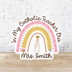 May include: A sticker with a rainbow design and the text "In My Catholic Teacher Era" in a circular layout. The rainbow features pink, yellow, and brown arches, with a depiction of a religious figure at the center. The name "Mrs. Smith" is at the bottom.