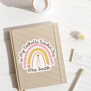 May include: A beige journal with a sticker featuring a rainbow design and the text "In My Catholic Teacher Era Mrs. Smith." The sticker includes a depiction of a religious figure and a crown. A white pen and a small notepad are also visible.