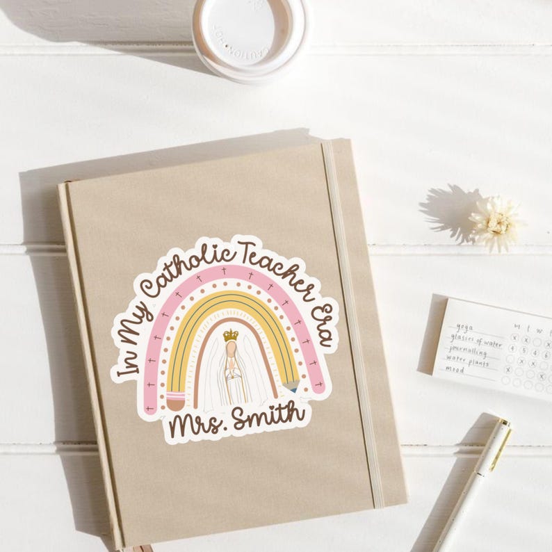 May include: A beige journal with a sticker featuring a rainbow design and the text "In My Catholic Teacher Era Mrs. Smith." The sticker includes a depiction of a religious figure and a crown. A white pen and a small notepad are also visible.