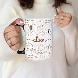Peut inclure: Mug en c&eacute;ramique blanche avec int&eacute;rieur et anse noirs, orn&eacute; de motifs religieux, d'&eacute;l&eacute;ments floraux et du pr&eacute;nom "alison" en &eacute;criture cursive. Le texte "Virgin Mary Pray for us" est &eacute;galement pr&eacute;sent.