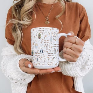 Peut inclure: Mug en c&eacute;ramique blanche avec un motif r&eacute;p&eacute;t&eacute; d'illustrations religieuses et le texte "Virgin Mary, Pray for us." Le motif comprend des images de la Vierge Marie, des &eacute;l&eacute;ments floraux et des accents dor&eacute;s. La tasse a une grande anse.