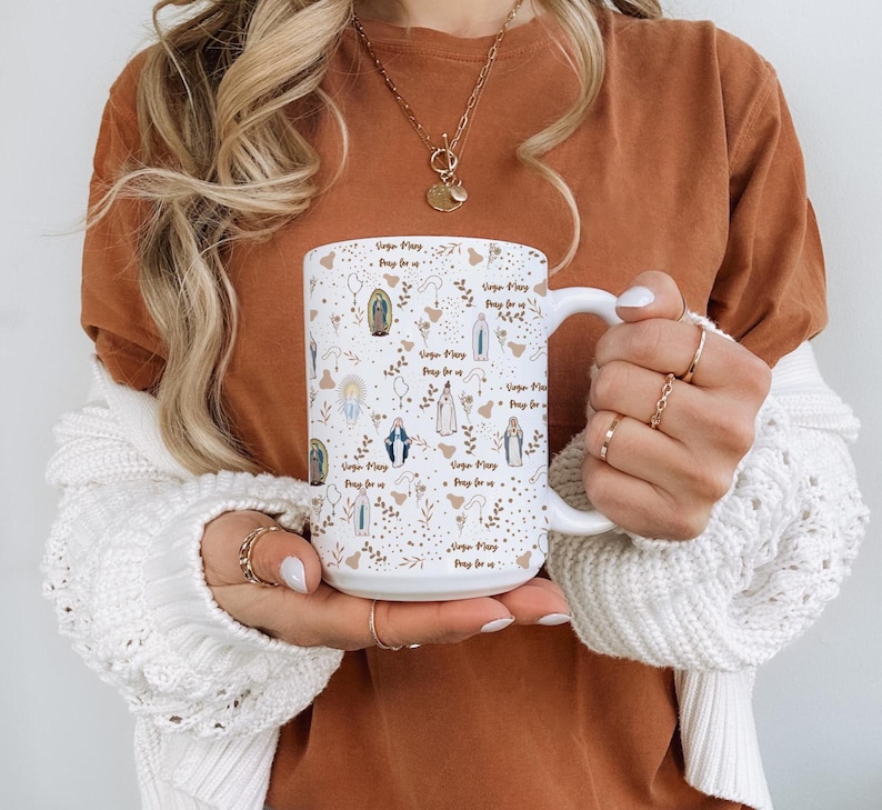 Peut inclure: Mug en c&eacute;ramique blanche avec un motif r&eacute;p&eacute;t&eacute; d'illustrations religieuses et le texte "Virgin Mary, Pray for us." Le motif comprend des images de la Vierge Marie, des &eacute;l&eacute;ments floraux et des accents dor&eacute;s. La tasse a une grande anse.
