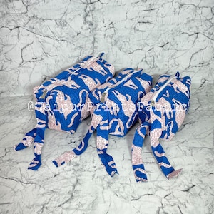 May include: Three blue and white patterned frog-shaped pillows with white zippers. The pillows have long, dangling legs and are arranged on a marble-patterned surface. The frog-shaped pillows are a unique home decor item.