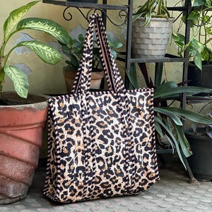 May include: A leopard print tote bag with black and white striped handles and trim. The bag has a quilted pattern and is made from a durable material. The bag is suitable for carrying various items and is a stylish accessory.