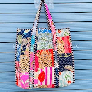 May include: A patchwork tote bag featuring a mix of vibrant fabric patterns, such as tiger stripes, floral motifs, and geometric shapes. The bag is trimmed with black and white stripes and has matching handles. It hangs against a blue backdrop.