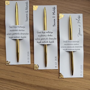 May include: Three gold pens with white cards. The cards have gold foil accents and text in Turkish, including names and a date. The pens are displayed on a wooden surface. The text includes the date 14.02.2026.