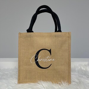 Könnte beinhalten: Eine naturbraune Jute-Tragetasche mit schwarzen Henkeln. Die Tasche hat einen großen schwarzen Buchstaben "C" und den Namen "Christine" in weißer Kursivschrift.