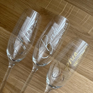 May include: Three clear champagne flutes with custom lettering. One says "Mrs. Jones" in silver, another says "Bride Vanessa" in silver, and the third says "Sophia Bridesmaid" in gold. The flutes are on a wooden surface.