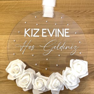 May include: A clear round acrylic sign with white lettering that reads "KIZ EVINE Hos Geldiniz" and is decorated with white flowers and white pearl-like embellishments.