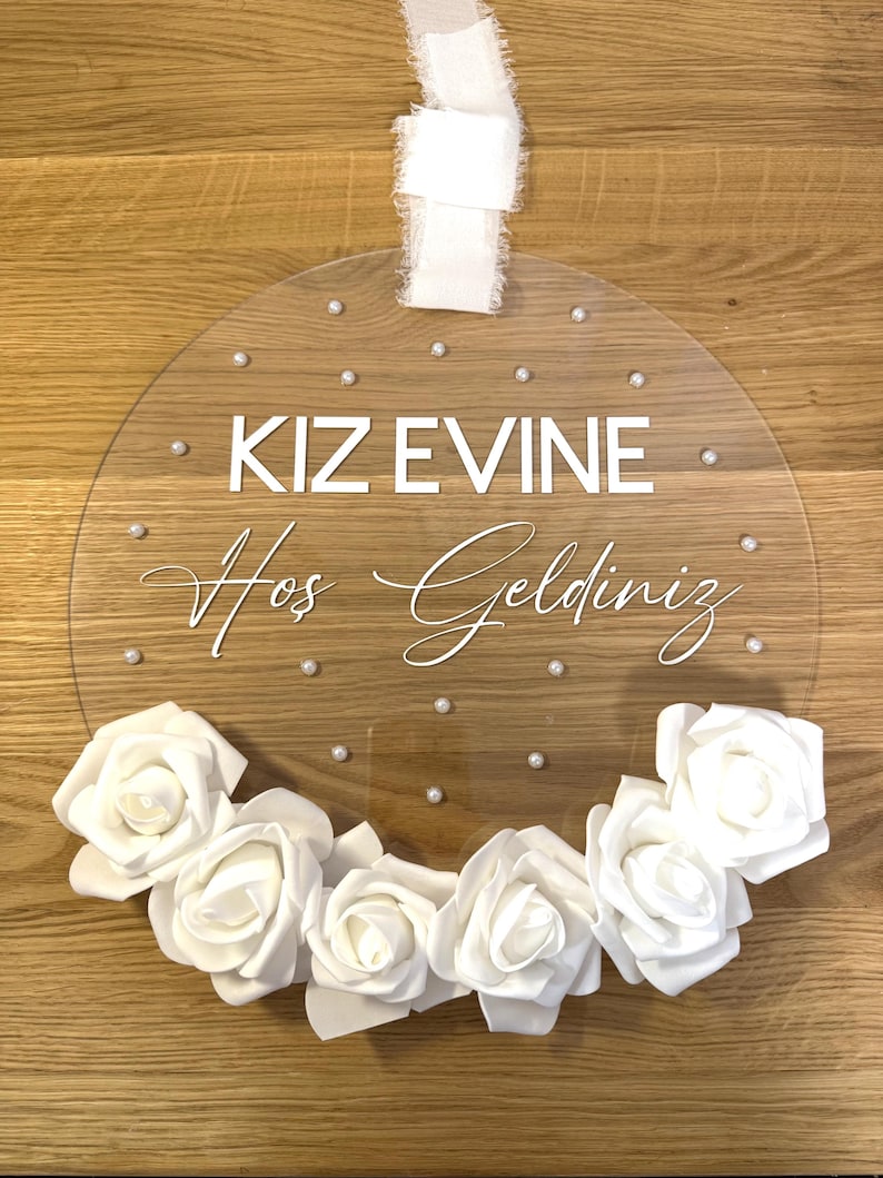May include: A clear round acrylic sign with white lettering that reads "KIZ EVINE Hos Geldiniz" and is decorated with white flowers and white pearl-like embellishments.
