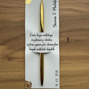 May include: A gold pen rests on a white card with gold foil accents. The card features the names "Yasemin & Mustafa" and the date "14. 02. 2026". Text in Turkish is also present on the card.