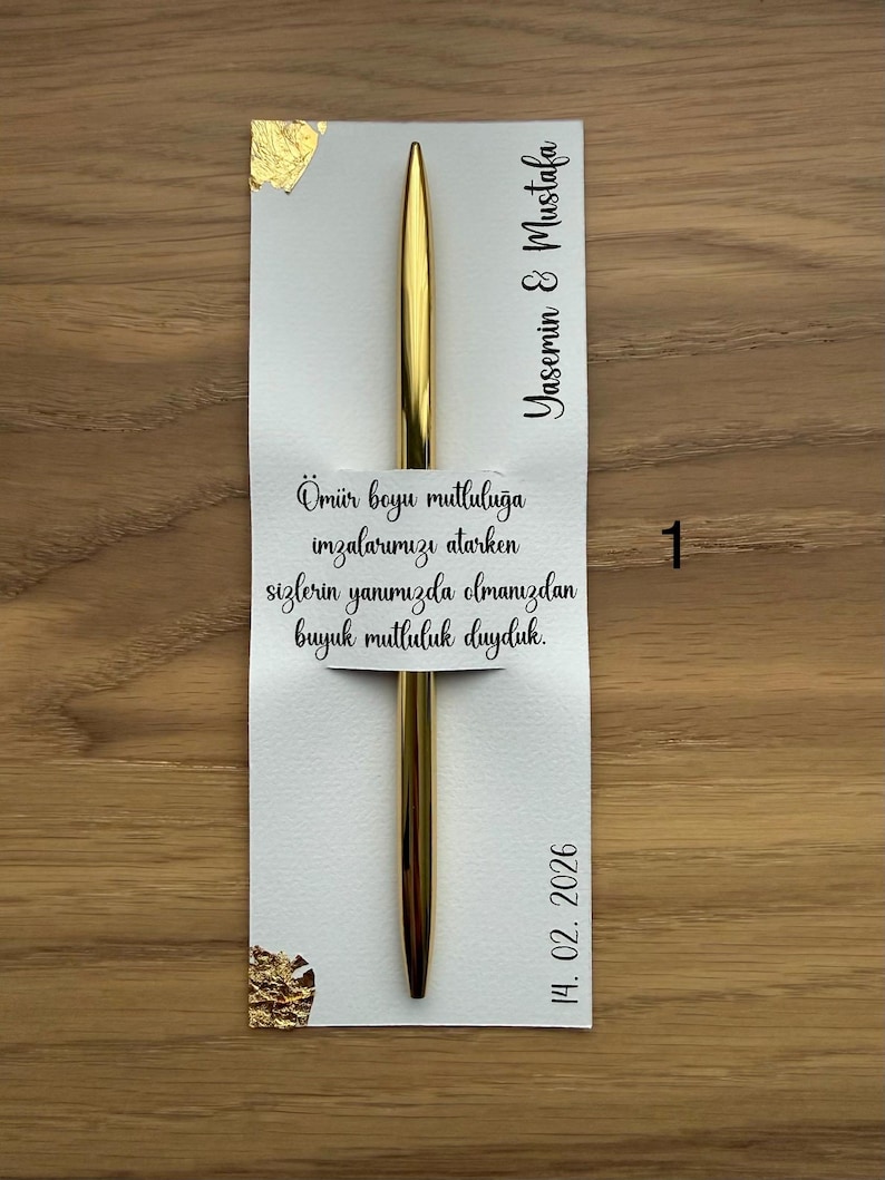 May include: A gold pen rests on a white card with gold foil accents. The card features the names "Yasemin & Mustafa" and the date "14. 02. 2026". Text in Turkish is also present on the card.