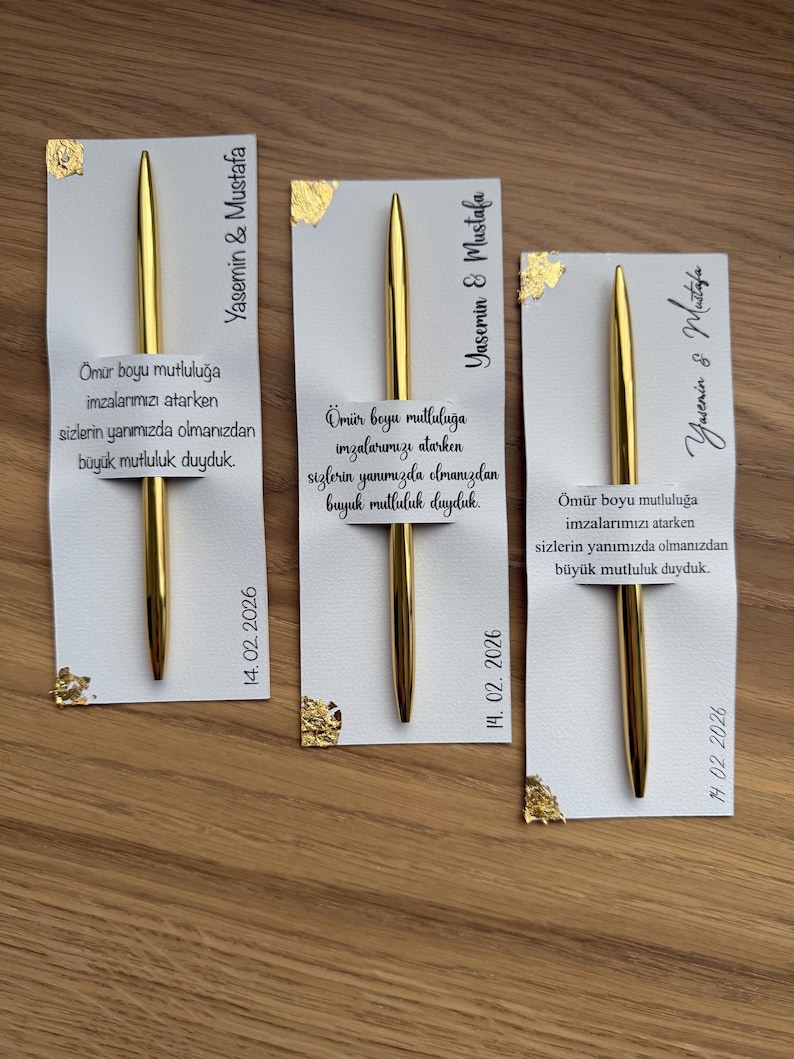 May include: Three gold pens with white cards. The cards have gold foil accents and text in Turkish, including names and a date. The pens are displayed on a wooden surface. The text includes the date 14.02.2026.