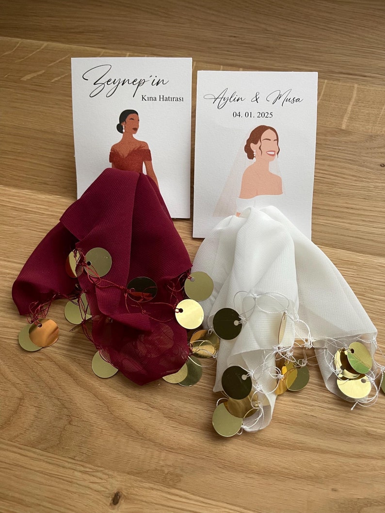 Op de afbeelding: Twee witte kaarten met illustraties van vrouwen in trouwjurken. De kaart links zegt "Zeynep'in Kına Hatırası". De kaart rechts zegt "Aylin & Musa 04.01.2025". De kaarten liggen op twee vierkante stukken stof, een rode en een witte, met gouden pailletten.