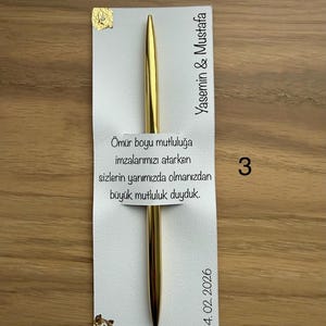 May include: A gold pen rests on a white card with Turkish text and the names Yasemin & Mustafa. The card also includes the date 14.02.2026 and the number 3. Gold leaf accents the card.