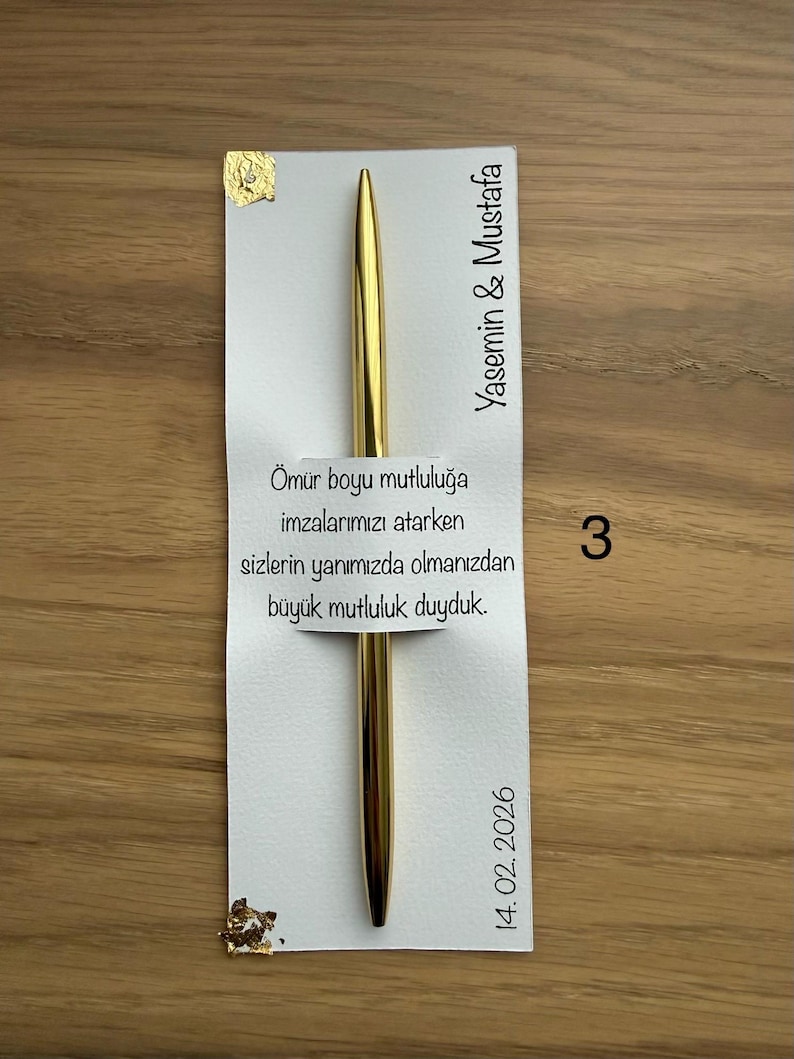 May include: A gold pen rests on a white card with Turkish text and the names Yasemin & Mustafa. The card also includes the date 14.02.2026 and the number 3. Gold leaf accents the card.