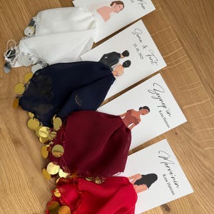 Op de afbeelding: Vier witte kaarten met kalligrafietekst en illustraties van paren. De kaarten zijn gelabeld met namen en data. De kaarten zijn in een rij op een houten oppervlak gerangschikt. Elke kaart is gekoppeld aan een klein, kleurrijk vierkant van stof met gouden munten die aan de randen zijn genaaid.