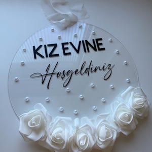 May include: White round acrylic sign with the text "KIZ EVINE Hosgeldiniz" in black lettering and white pearl accents. The sign is decorated with a white flower garland.
