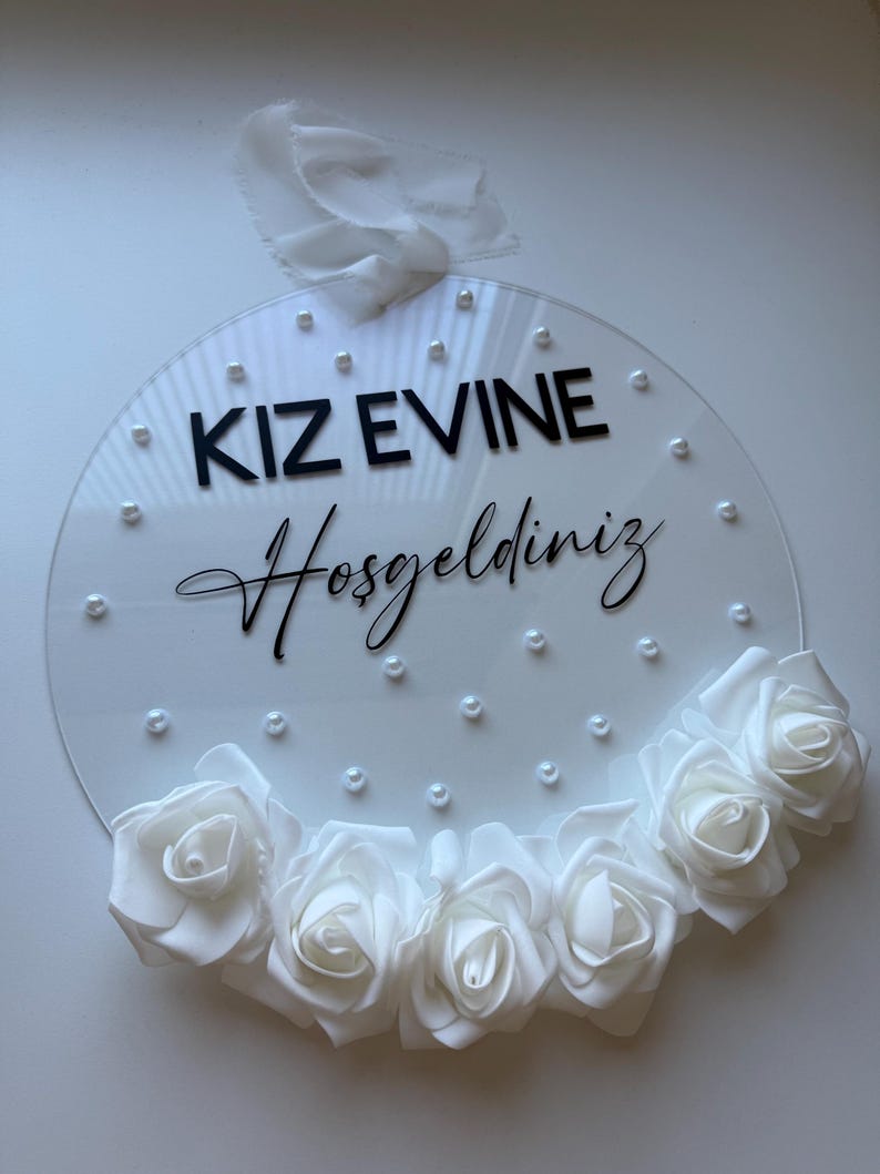May include: White round acrylic sign with the text "KIZ EVINE Hosgeldiniz" in black lettering and white pearl accents. The sign is decorated with a white flower garland.