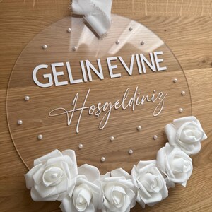 May include: A clear round acrylic sign with the text "GELIN EVINE Hosgeldiniz" in white lettering. The sign is decorated with white pearl-like beads and a garland of white roses.