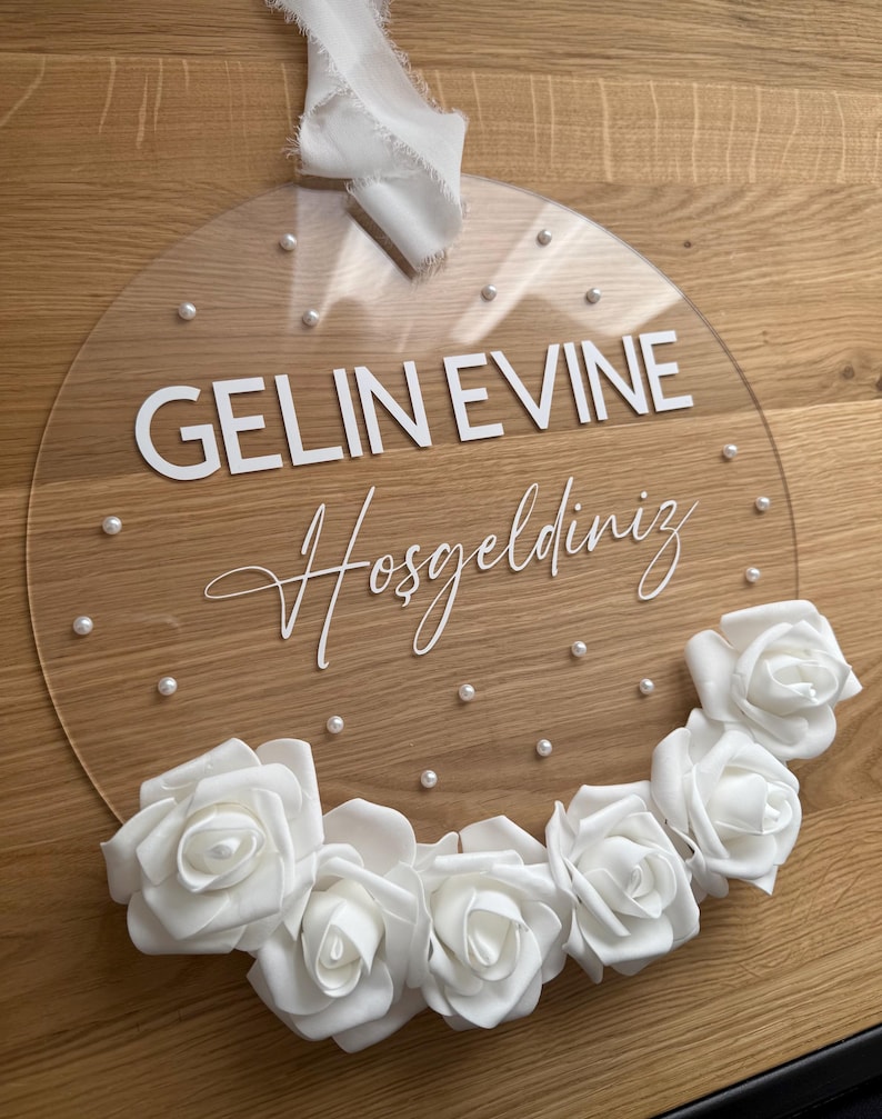 May include: A clear round acrylic sign with the text "GELIN EVINE Hosgeldiniz" in white lettering. The sign is decorated with white pearl-like beads and a garland of white roses.