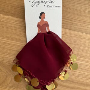 Op de afbeelding: Een bordeauxrode chiffon sjaal met gouden muntversieringen. De sjaal is in een driehoekige vorm gevouwen en heeft een decoratieve franje. De tekst "Zeynep'in Kına Hatırası" is gedrukt op een witte kaart achter de sjaal.