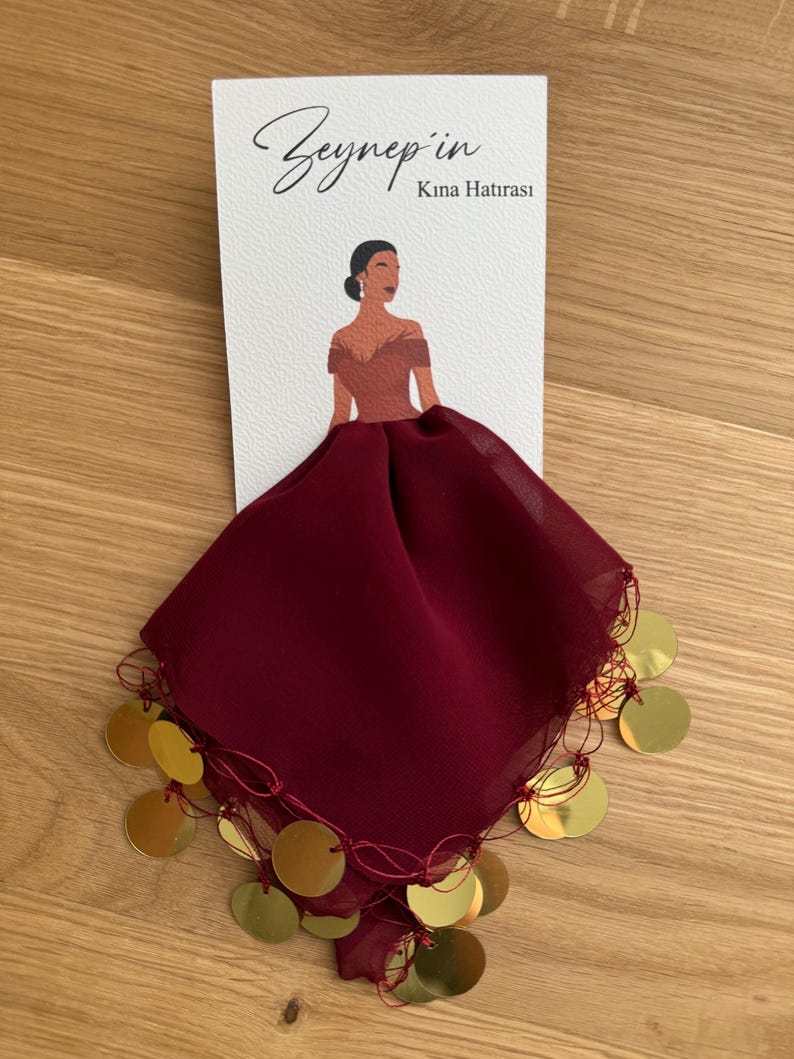 Op de afbeelding: Een bordeauxrode chiffon sjaal met gouden muntversieringen. De sjaal is in een driehoekige vorm gevouwen en heeft een decoratieve franje. De tekst "Zeynep'in Kına Hatırası" is gedrukt op een witte kaart achter de sjaal.