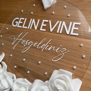 May include: A clear round acrylic sign with the text "GELIN EVINE Hosgeldiniz" in white lettering. The sign is decorated with white pearl-like embellishments and white roses.