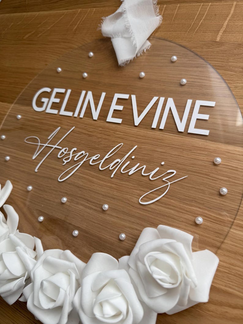 May include: A clear round acrylic sign with the text "GELIN EVINE Hosgeldiniz" in white lettering. The sign is decorated with white pearl-like embellishments and white roses.