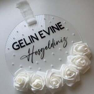 May include: White round acrylic sign with the text "GELIN EVINE Hosgeldiniz" in black lettering and a white floral garland around the edge.