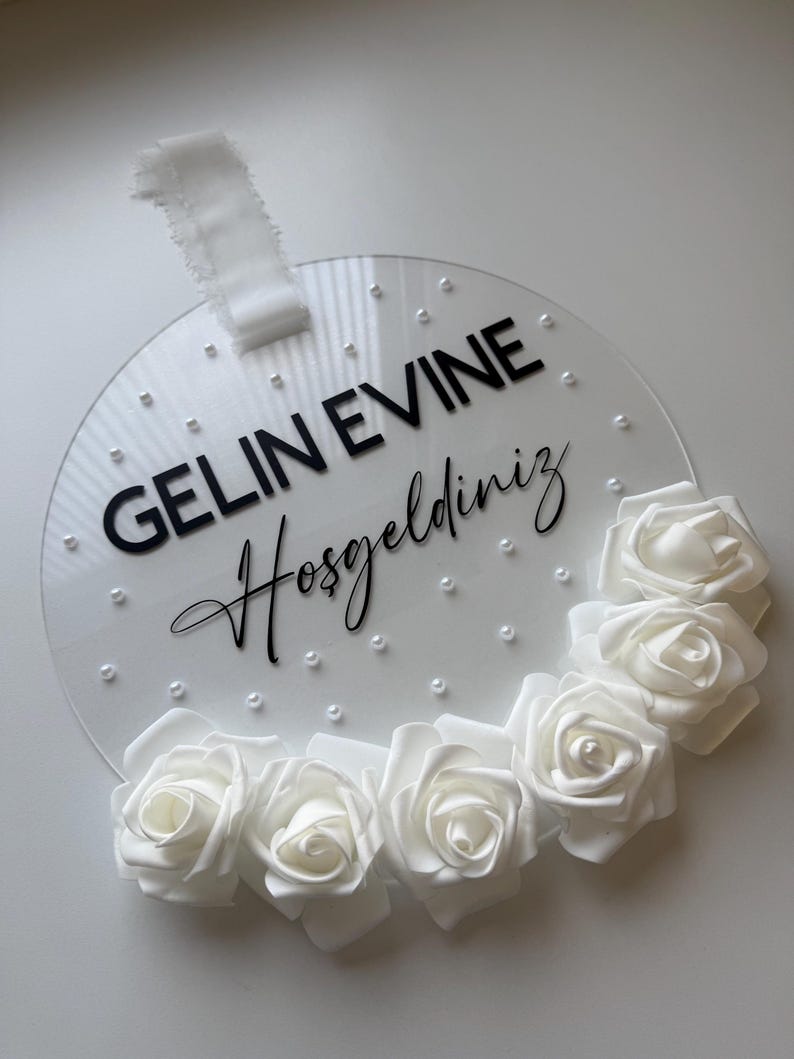 May include: White round acrylic sign with the text "GELIN EVINE Hosgeldiniz" in black lettering and a white floral garland around the edge.