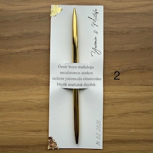 May include: A gold pen rests on a white card with gold leaf accents. The card has text in a script font and the date 14.02.2026. The background is a light brown wooden surface.