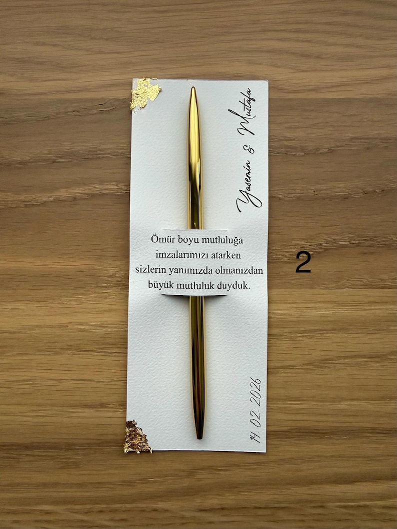 May include: A gold pen rests on a white card with gold leaf accents. The card has text in a script font and the date 14.02.2026. The background is a light brown wooden surface.