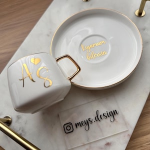May include: A white ceramic teacup and saucer set with gold trim and lettering, resting on a marble tray with gold handles. The teacup features gold initials and a heart. The saucer has gold text. A clear label with the Instagram handle "meys.design" is also present.