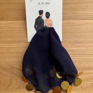 Op de afbeelding: Een witte kaart met de tekst "Sema & Ferit 25.05.2025" en een aquarel-illustratie van een stel in trouwkledij. De kaart is bevestigd aan een donkerblauwe stof met gouden pailletten.