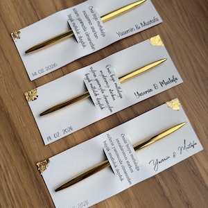 May include: Three white cards with gold foil accents and gold pens. Each card has text and the date 14.02.2026. The names "Yasemin & Mustafa" are written on each card.