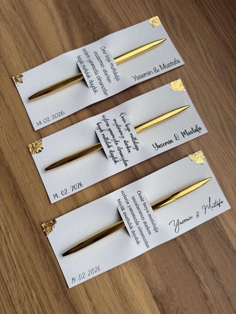 May include: Three white cards with gold foil accents and gold pens. Each card has text and the date 14.02.2026. The names "Yasemin & Mustafa" are written on each card.