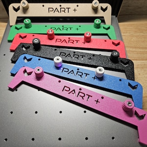 May include: A collection of colorful metal tools, each with the text "PART +" and adjustable knobs. The tools are in beige, green, red, black, blue, and pink, arranged on a gray surface with a wooden background.