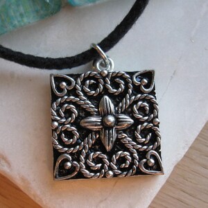 May include: Silver pendant necklace with a square-shaped charm featuring an intricate design of interlacing ropes and a central flower motif.