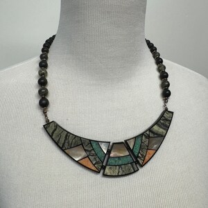 May include: A black and green stone necklace with a geometric pendant. The pendant is made of various stones, including green, black, and white.