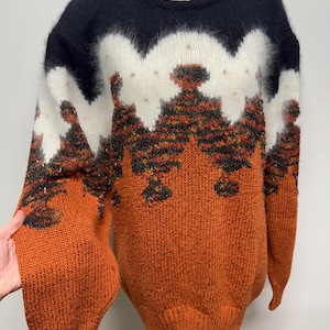 Puede incluir: Un suéter oversize en tonos naranja, negro y blanco, con un diseño inspirado en un bosque y adornos de perlas. El suéter de cuello redondo tiene mangas largas. Una prenda cálida y elegante. El diseño evoca una escena de bosque.