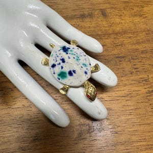 May include: A white ceramic turtle brooch with blue and green splatter paint accents. The turtle has gold accents and red eyes.