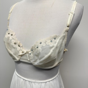 May include: Ivory-colored bra with floral embroidery along the neckline. The bra features adjustable straps and a delicate bow detail. The bra is paired with a matching white slip.