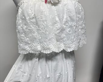 Puede incluir: Vestido blanco de ojal con top sin tirantes y falda amplia. El vestido presenta intrincados bordados florales y un dobladillo festoneado. Una flor de tela blanca con un acento de cinta rosa está prendida en la parte superior.